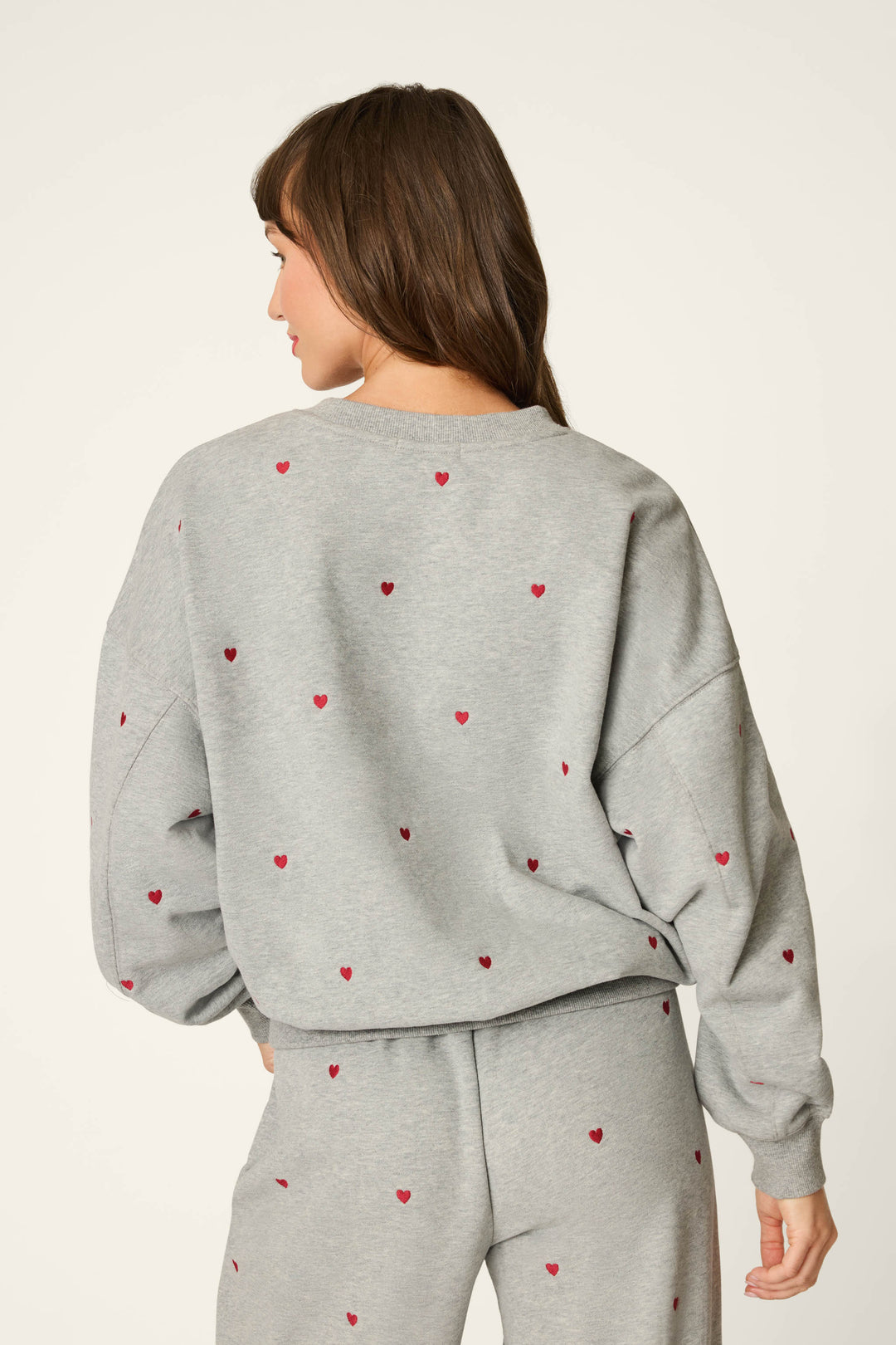A woman with long brown hair faces away, wearing the P.J. Salvage C'est La Vie Embroidered Hearts Frankie Sweatshirt and matching pants adorned with small red hearts.