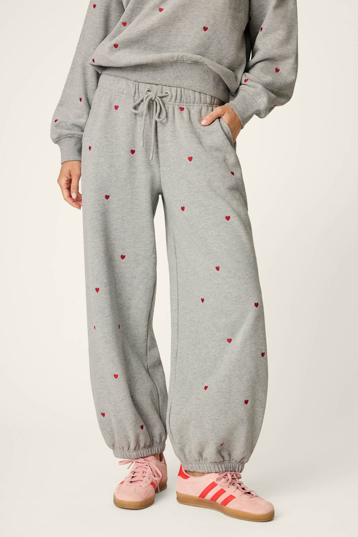 A person is shown from the shoulders down wearing P.J. Salvage's C'est La Vie Embroidered Hearts Good Hang Sweatpants, featuring small red heart prints, paired with pink Adidas sneakers with red stripes. One hand rests in their pocket.