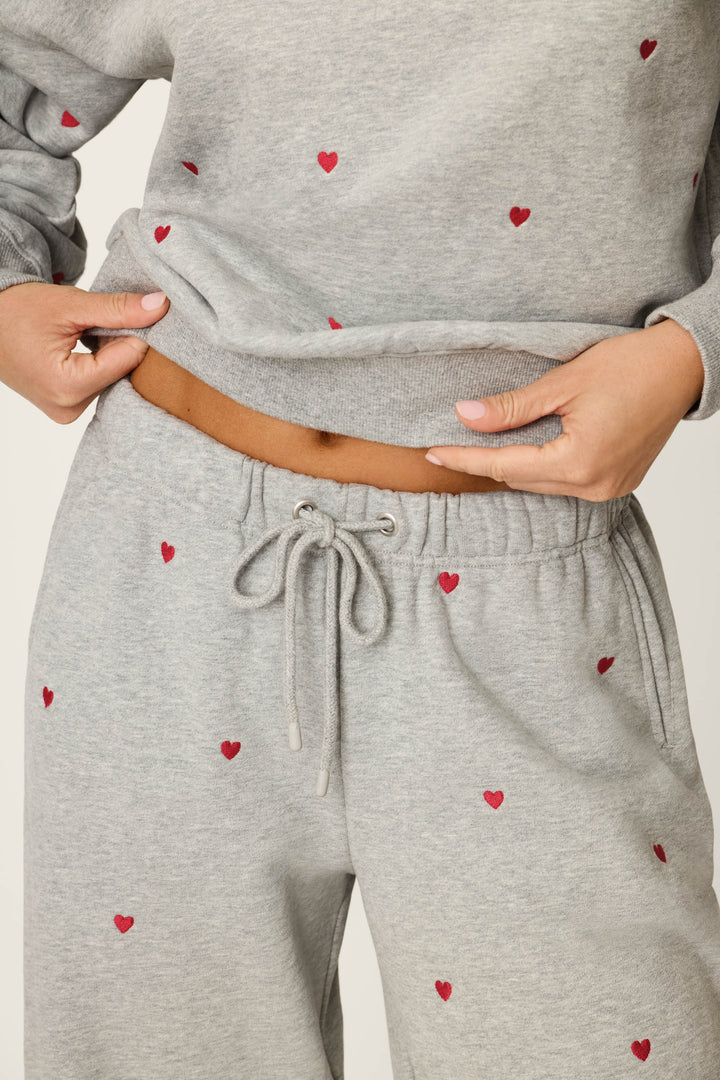 A person wearing P.J. Salvage's C'est La Vie Embroidered Hearts Good Hang Sweatpants, featuring small red heart patterns, pairs them with a light gray sweatshirt and slightly lifts the shirt to reveal part of their midriff.
