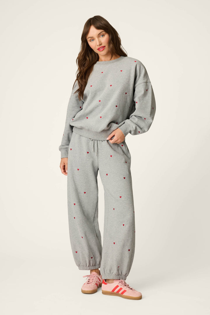A woman stands wearing the P.J. Salvage C'est La Vie Embroidered Hearts Good Hang Sweatpants in a relaxed grey fit, featuring small red hearts, paired with a matching sweatshirt and pink and red sneakers. She poses casually against a plain, light background.