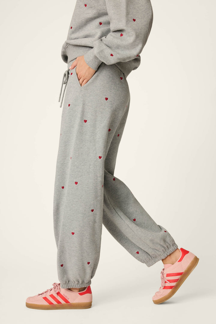 A person wearing the P.J. Salvage C'est La Vie Embroidered Hearts Good Hang Sweatpants and a matching grey sweatshirt featuring small red heart prints, paired with pink Adidas sneakers with red stripes, is shown from the side walking with one hand in their pocket.