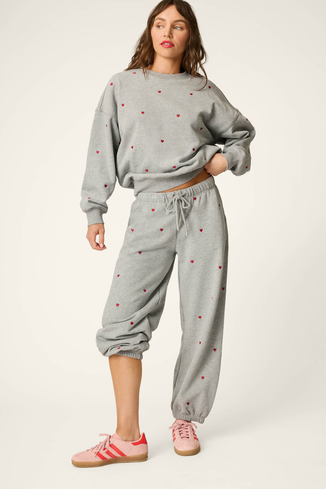 A woman with brown hair stands against a plain light background, wearing P.J. Salvage's C'est La Vie Embroidered Hearts Good Hang Sweatpants—a loose grey pair featuring small pink heart prints, with one pant leg pulled up. She completes the look with a matching sweatshirt and pink sneakers.