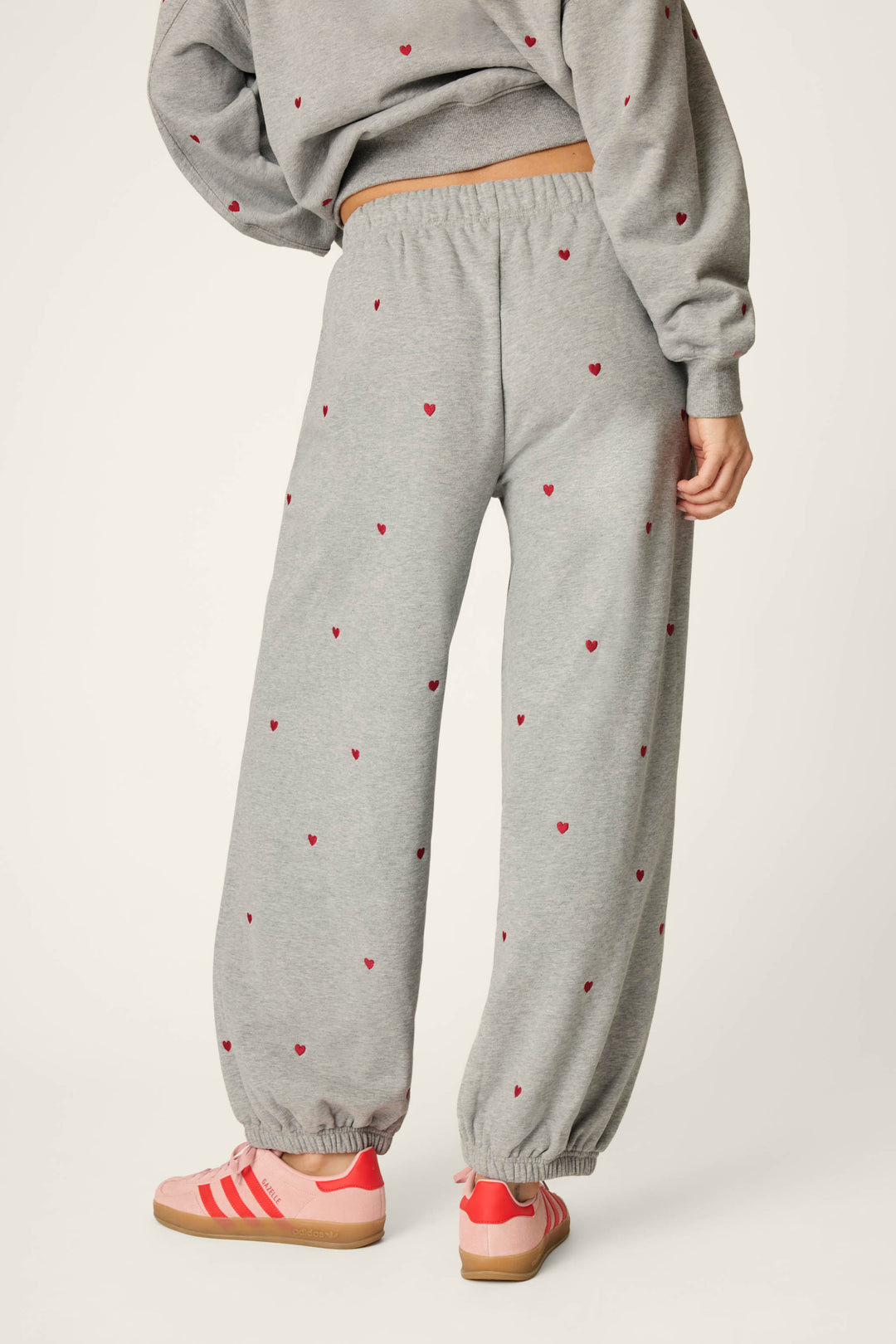 A person wearing the P.J. Salvage C'est La Vie Embroidered Hearts Good Hang Sweatpants—gray with small red heart details—paired with a matching gray sweatshirt and pink-and-red sneakers, stands against a light background. The photo is taken from behind to showcase the relaxed fit of the sweatpants.
