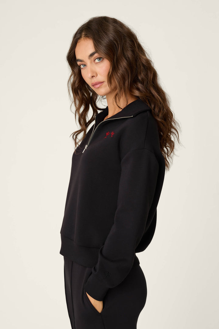 A woman modeling a black half-zip sweater with a small red embroidered logo on the left chest, paired with black pants, against a plain light background.
