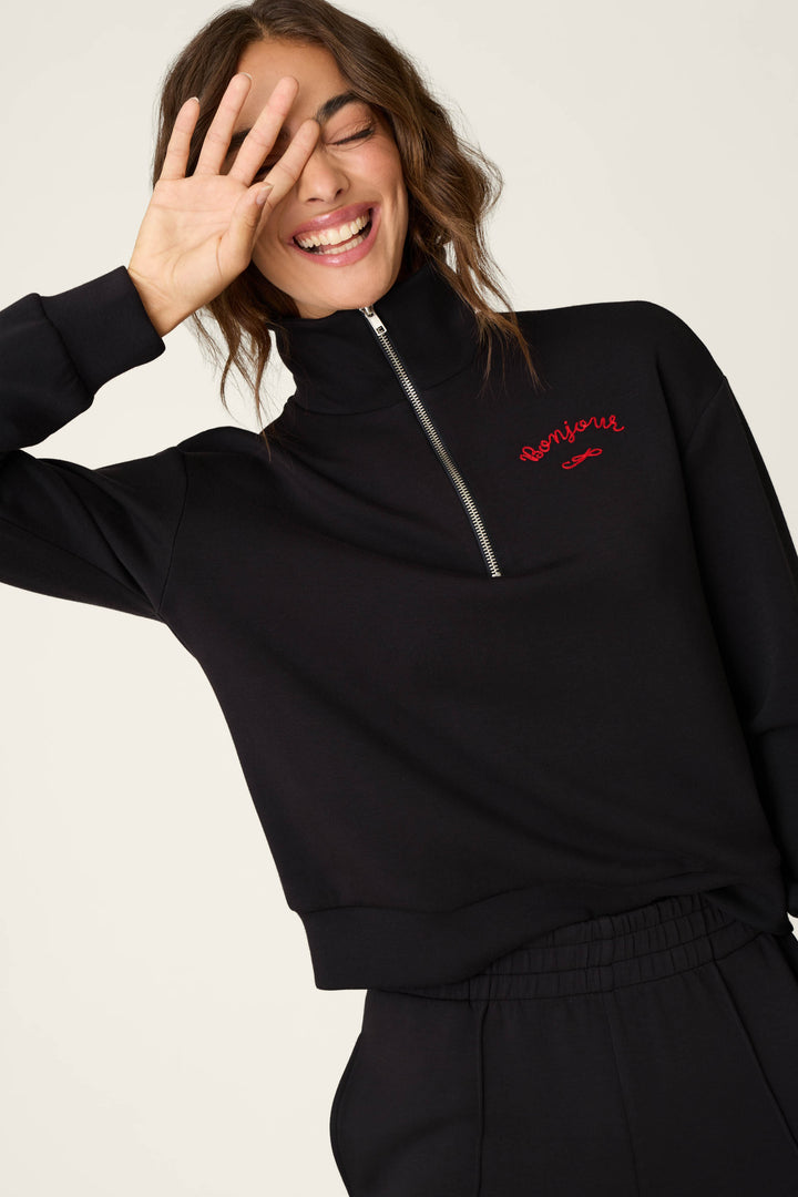 A woman smiles with her eyes closed, raising one hand near her face, while wearing the P.J. Salvage C'est La Vie Midnight Flights Overnight Sweater—a black zip-up top embroidered with "Bonjour" in red.