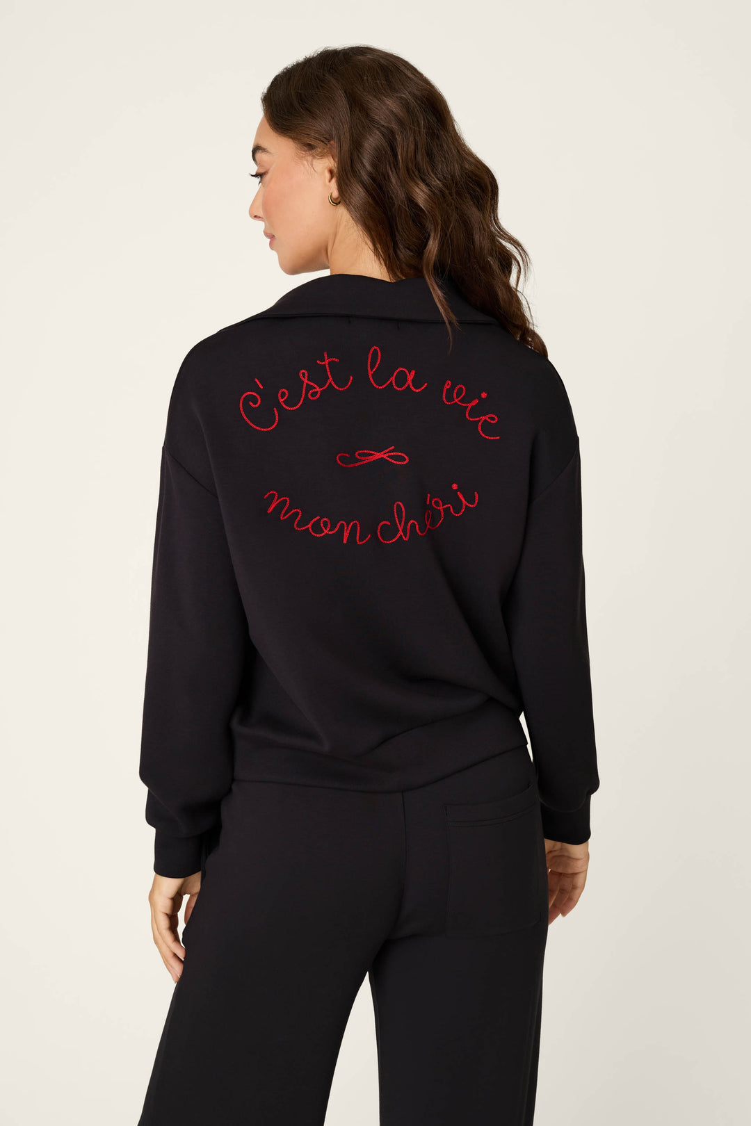 A woman shown from the back wearing a sweater with red cursive text reading 'C'est la vie mon chéri' embroidered across the back, paired with black pants, against a plain light background.