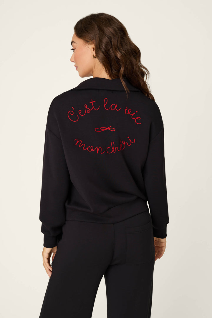A woman shown from the back wearing a sweater with red cursive text reading 'C'est la vie mon chéri' embroidered across the back, paired with black pants, against a plain light background.