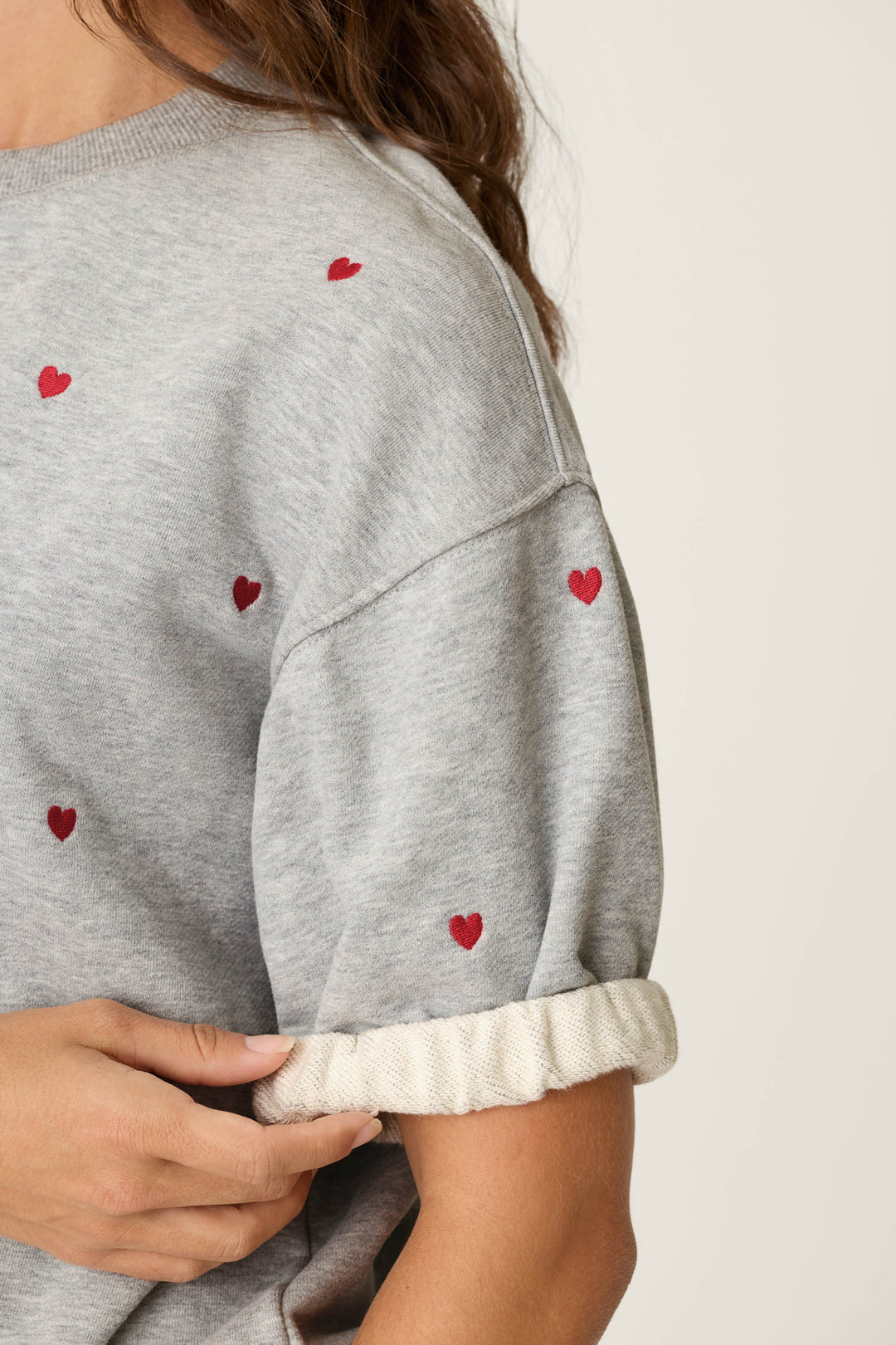 A person is seen from the torso up, wearing the P.J. Salvage C'est La Vie Embroidered Hearts Roll Back Short Sleeve Sweatshirt in light gray, featuring small red heart patterns. They are rolling up the cuff of one sleeve, with only their torso, part of their arm, and some hair visible against a plain background.