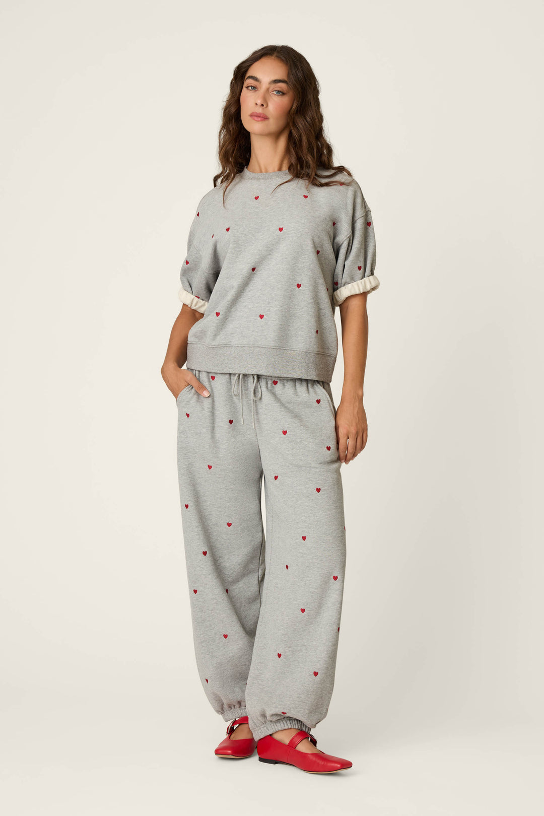 A woman wears the P.J. Salvage "C'est La Vie Embroidered Hearts" Roll Back Short Sleeve Sweatshirt in grey, paired with matching sweatpants featuring subtle red embroidered hearts. She completes the look with red flats and stands against a plain light background, one hand casually in her pocket.