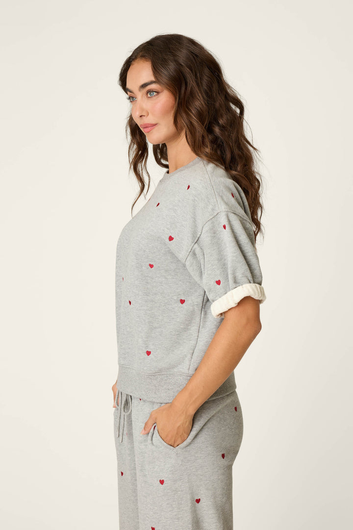 A woman with wavy brown hair stands in profile with her hands in her pockets against a plain background, wearing the P.J. Salvage C'est La Vie Embroidered Hearts Roll Back Short Sleeve Sweatshirt and matching pants featuring small red heart patterns.