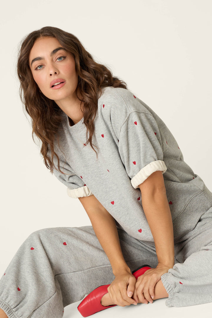 A woman with wavy brown hair is seated on the floor against a plain background, holding a red shoe and looking at the camera with a neutral expression. She wears the P.J. Salvage C'est La Vie Embroidered Hearts Roll Back Short Sleeve Sweatshirt, featuring a grey lounge style adorned with small red heart embroidery.