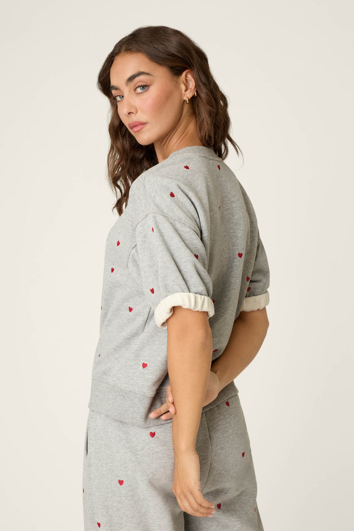 A woman with wavy brown hair stands sideways and looks over her shoulder, wearing the P.J. Salvage C'est La Vie Embroidered Hearts Roll Back Short Sleeve Sweatshirt and matching pants adorned with small red heart prints. The background is a plain off-white.