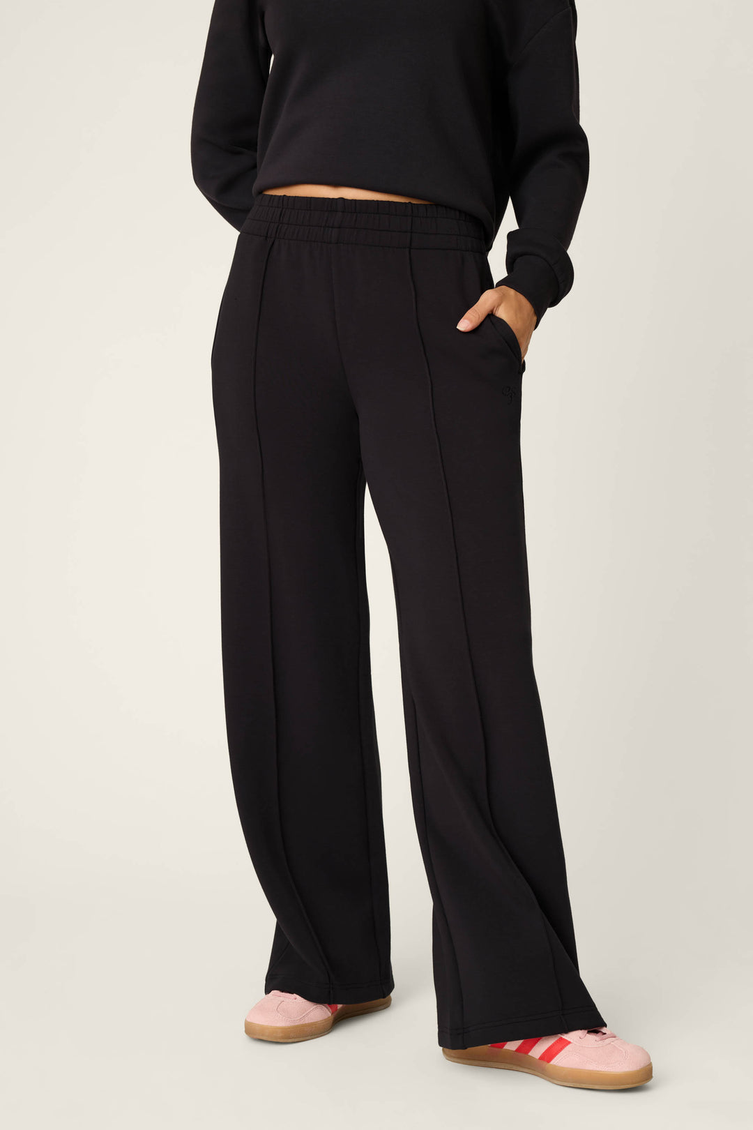 Wearing the C'est La Vie Midnight Flights Runway Pants by P.J. Salvage, this person stands with one hand in their pocket, styled with a black cropped sweatshirt and pink sneakers featuring red stripes against a plain background.