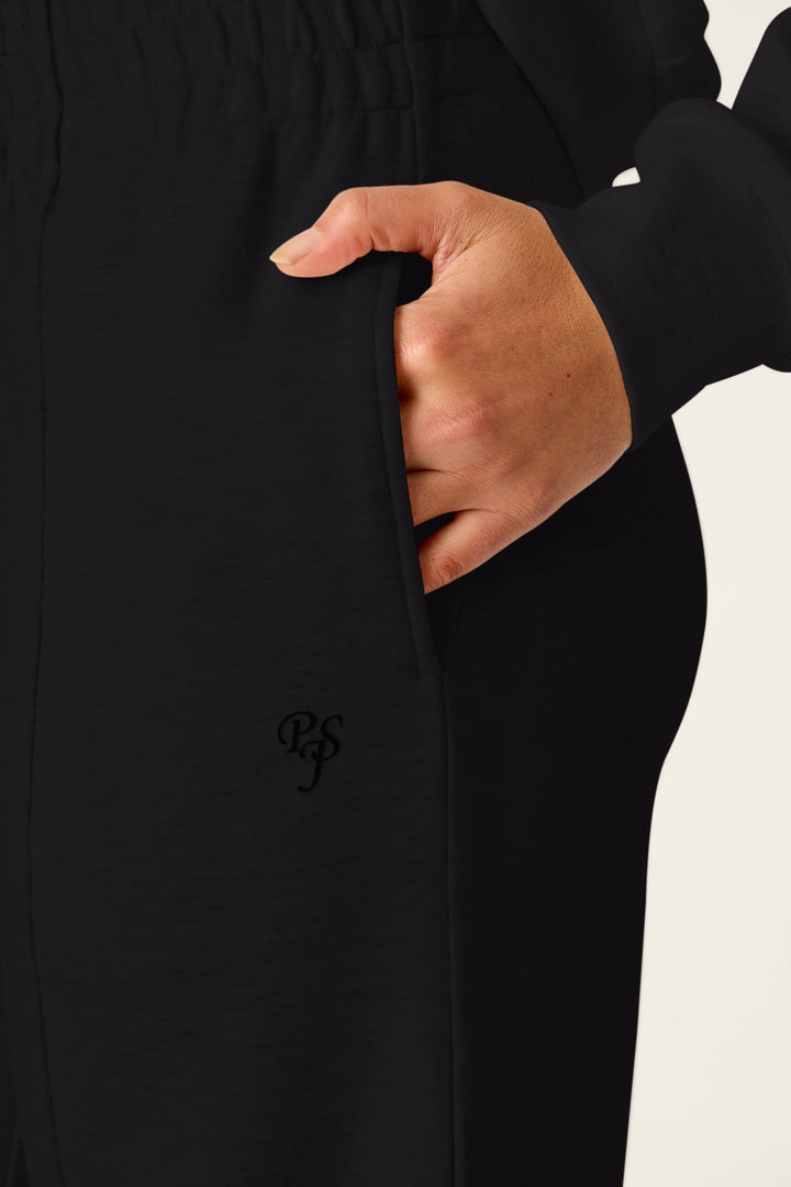 A close-up of a light-skinned hand slipping into the pocket of P.J. Salvage’s C'est La Vie Midnight Flights Runway Pants, featuring an embroidered PS logo, set against a plain background.
