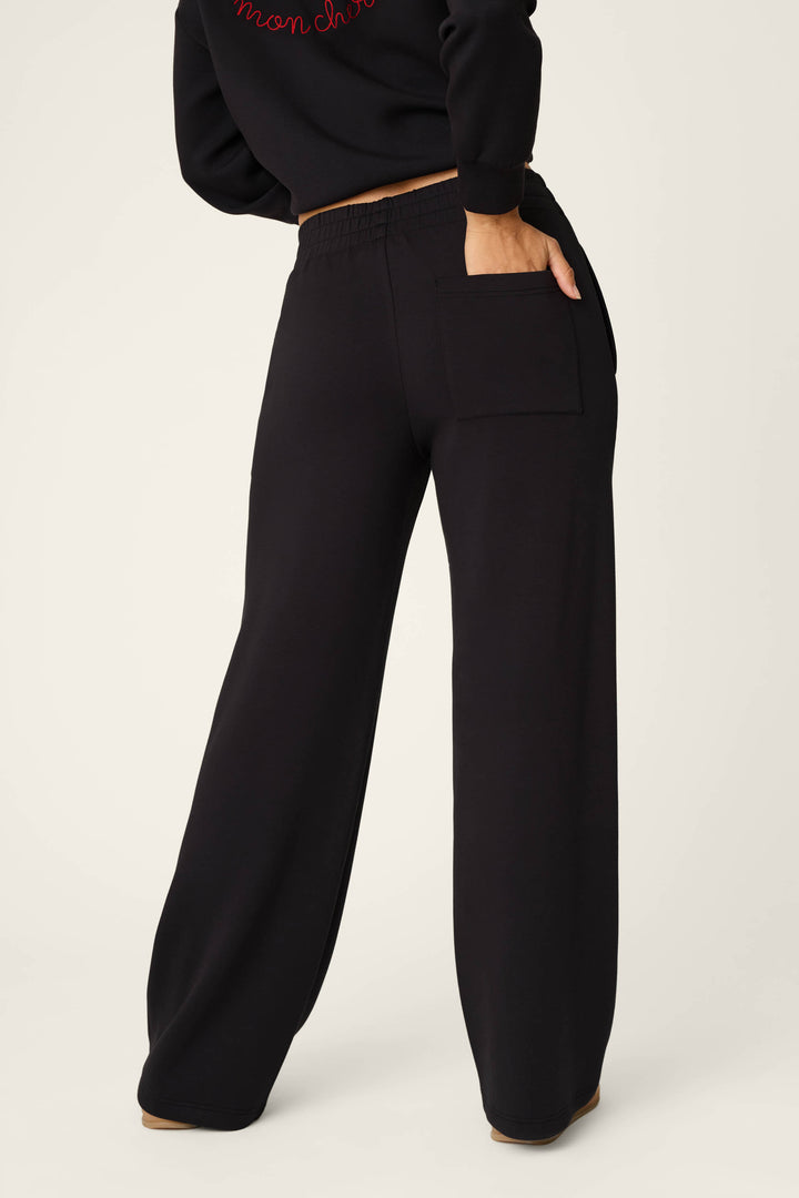 A person is seen from behind, standing with one hand in the pocket, wearing the P.J. Salvage C'est La Vie Midnight Flights Runway Pants—loose black pants featuring a single back pocket—paired with a black sweatshirt against a plain background.