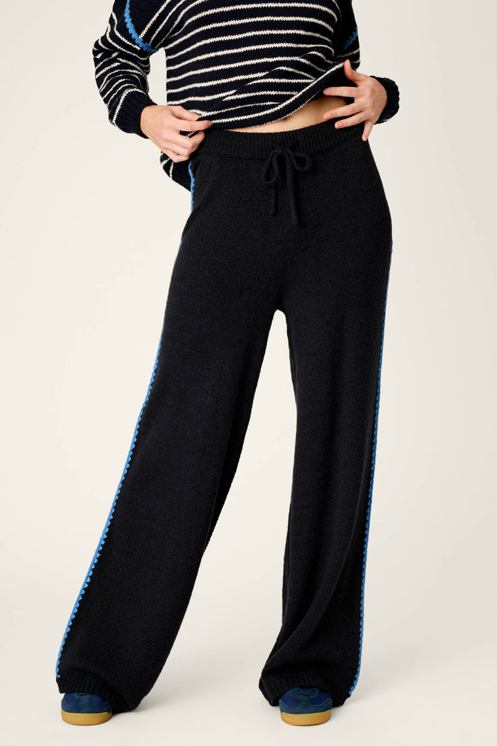 A person is shown from the shoulders down, hands on waistband, wearing P.J. Salvage C'est La Vie Suki Jogger Pants—black wide-leg joggers with a blue stripe down the sides—paired with a black and white striped sweater and blue sneakers.