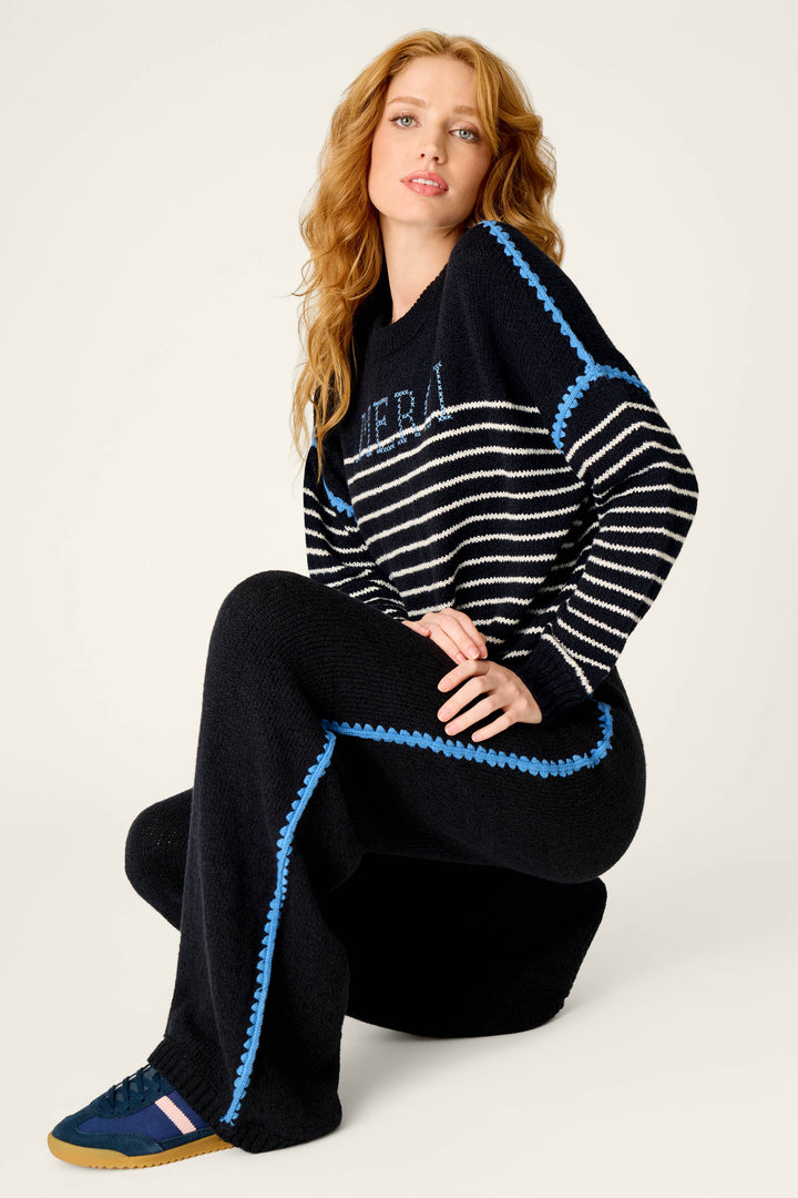 A woman with long wavy hair poses seated, wearing the C'est La Vie Suki Jogger Pants by P.J. Salvage, paired with a navy sweater featuring white stripes and blue trim, and navy sneakers with blue and pink details.