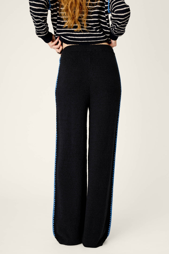A person stands with their back to the camera, wearing P.J. Salvage's C'est La Vie Suki Jogger Pants, which feature a black wide-leg design with a blue stripe down the side, paired with a black striped sweater. Their hands rest on their hips and their long, wavy hair is visible.