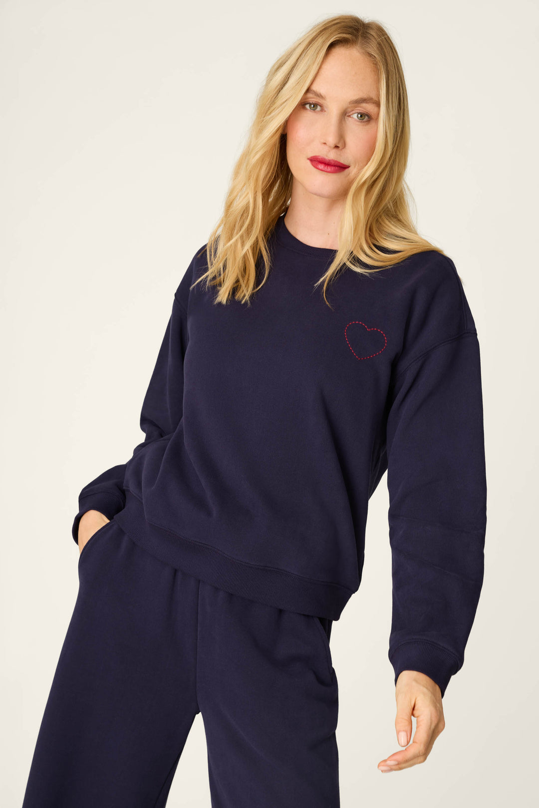 A woman with blonde hair and a slight smile stands with one hand in her pocket, wearing the P.J. Salvage C'est La Vie Blue Ink Simone Sweater, which features a small red stitched heart detail on the chest. She pairs the sweater with matching pants.