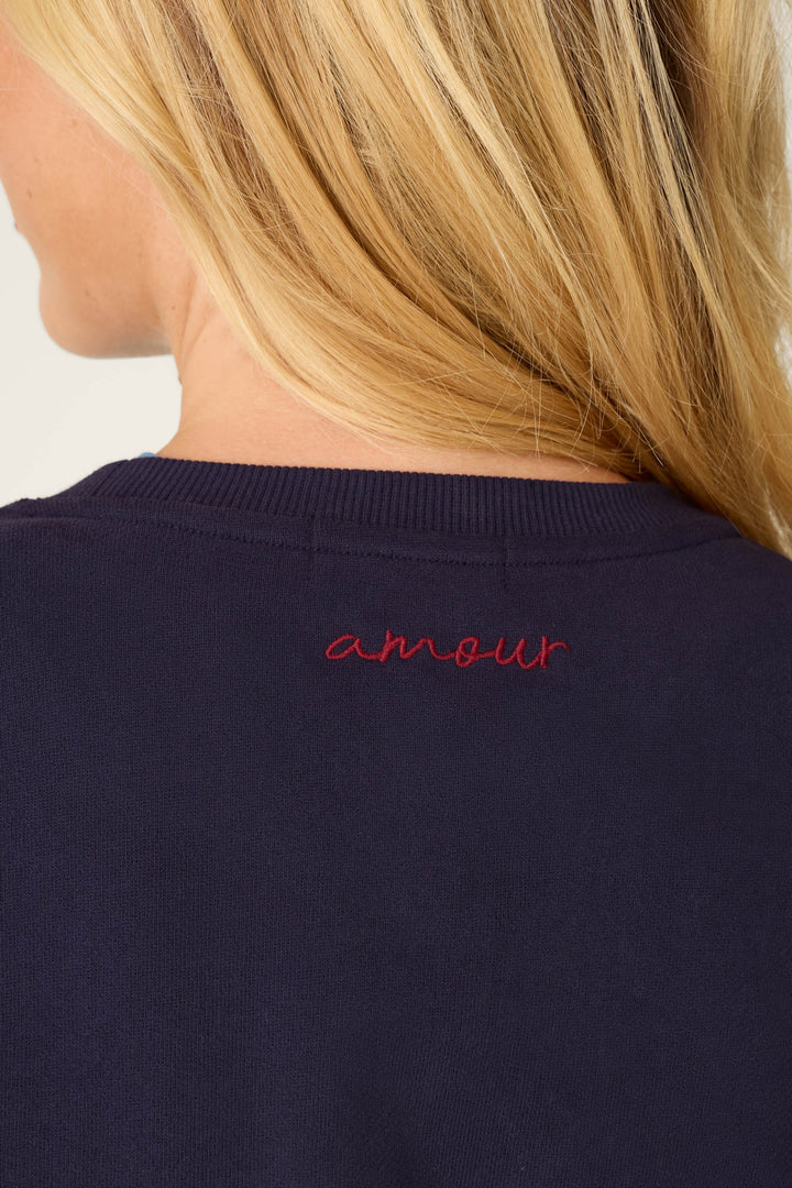A close-up of a person's back shows the word "amour" embroidered in red on the navy blue collar of the C'est La Vie Blue Ink Simone Sweater by P.J. Salvage. The person has long blonde hair.