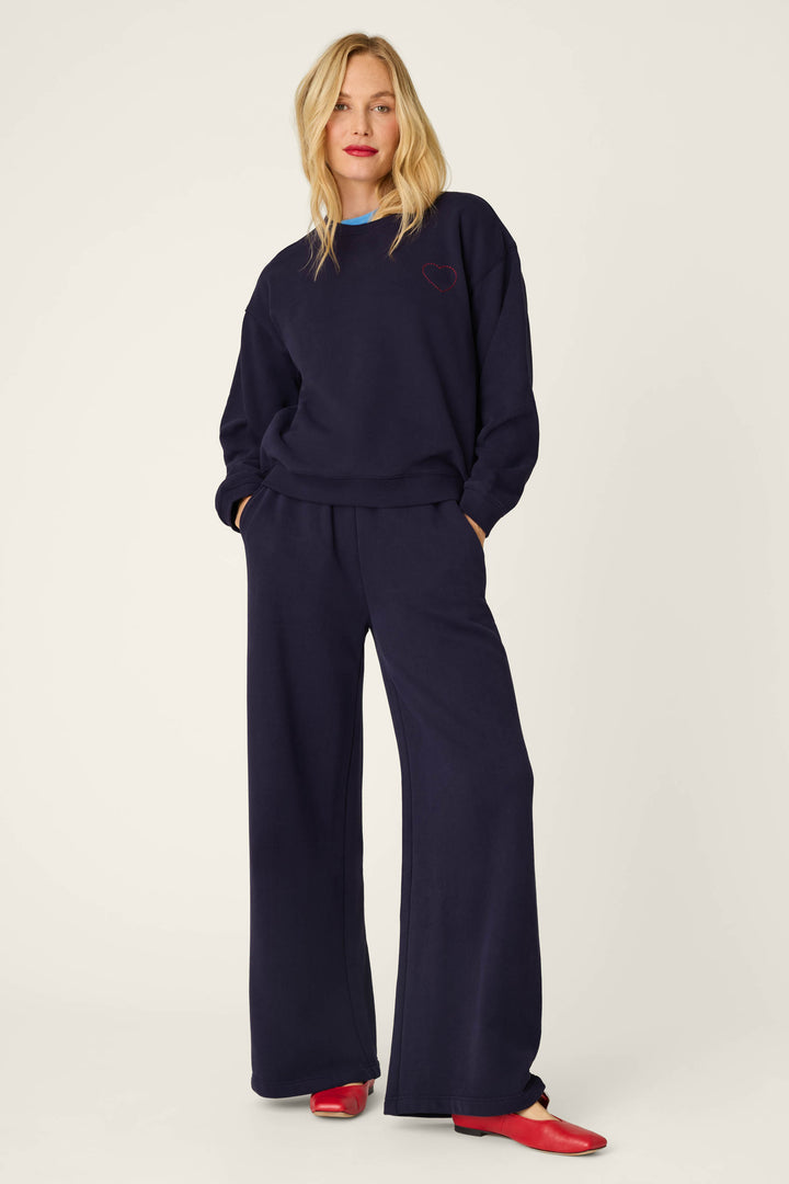 A woman stands with her hands in her pockets, wearing the P.J. Salvage C'est La Vie Blue Ink Simone Sweater paired with matching wide-leg pants. She has blonde hair and red shoes, and the sweater features a small heart detail. The background is plain off-white.