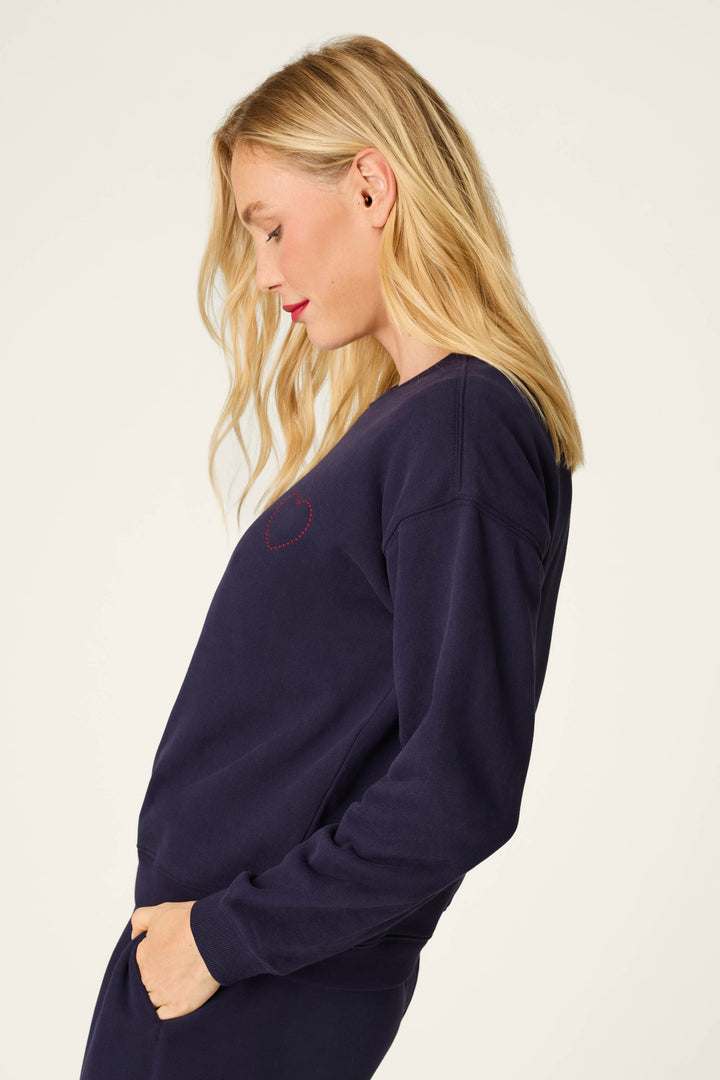 A woman with blonde hair stands in profile against a plain light background, wearing the P.J. Salvage "C'est La Vie Blue Ink Simone Sweater," a navy blue sweatshirt featuring a small red heart outline.