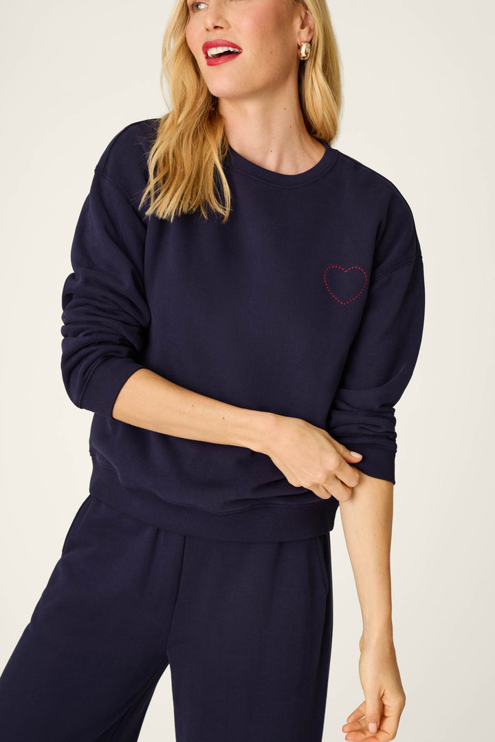 A woman is smiling with her head turned slightly to the side, wearing the P.J. Salvage C'est La Vie Blue Ink Simone Sweater featuring a small red heart embroidered on the left chest, paired with matching pants.