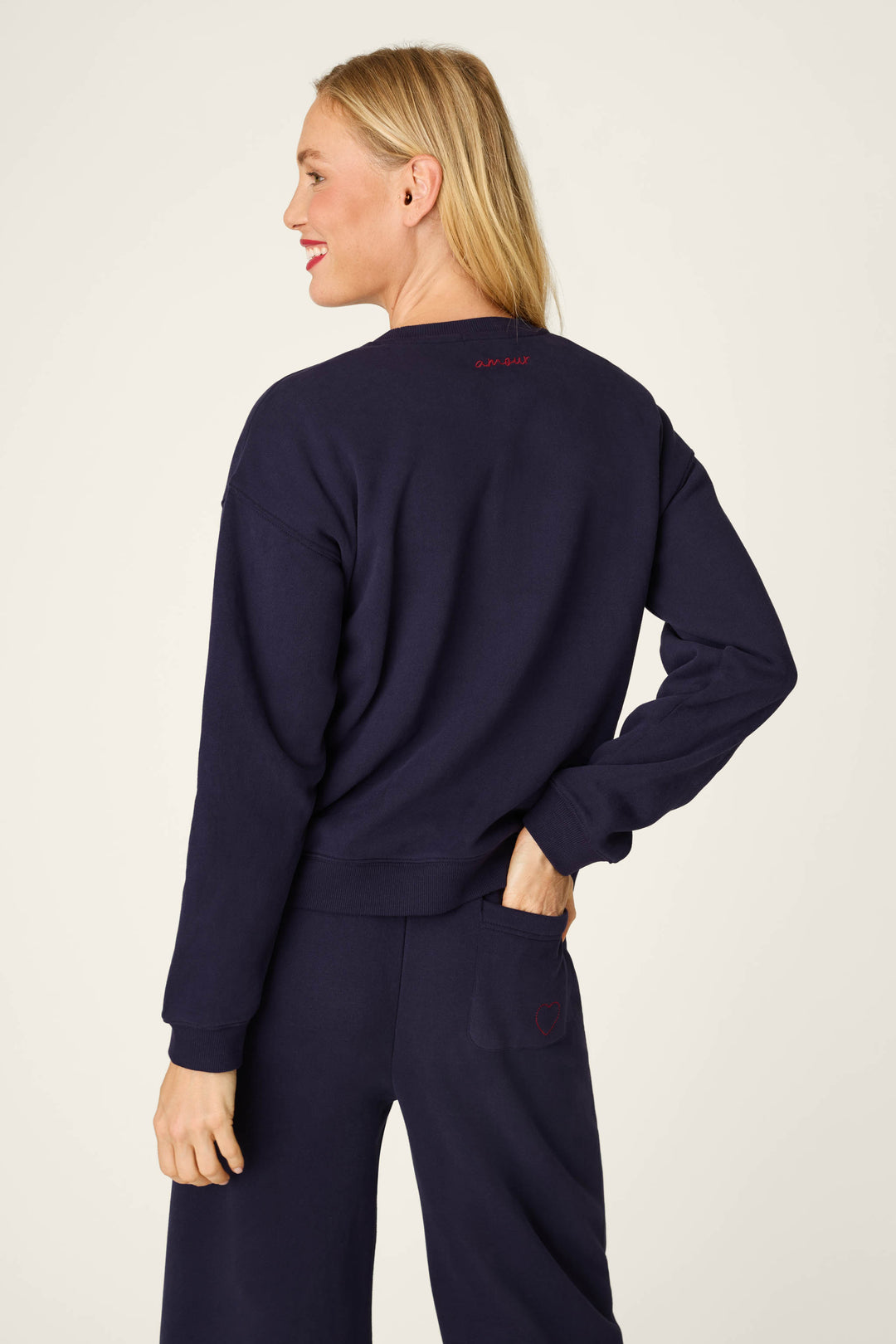 Wearing the P.J. Salvage C'est La Vie Blue Ink Simone Sweater and matching pants, a blonde-haired woman stands against a plain background, smiling over her shoulder with one hand in her back pocket.