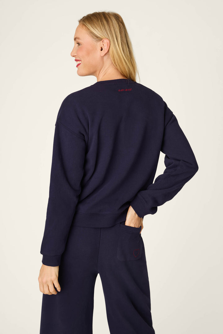 Wearing the P.J. Salvage C'est La Vie Blue Ink Simone Sweater and matching pants, a blonde-haired woman stands against a plain background, smiling over her shoulder with one hand in her back pocket.