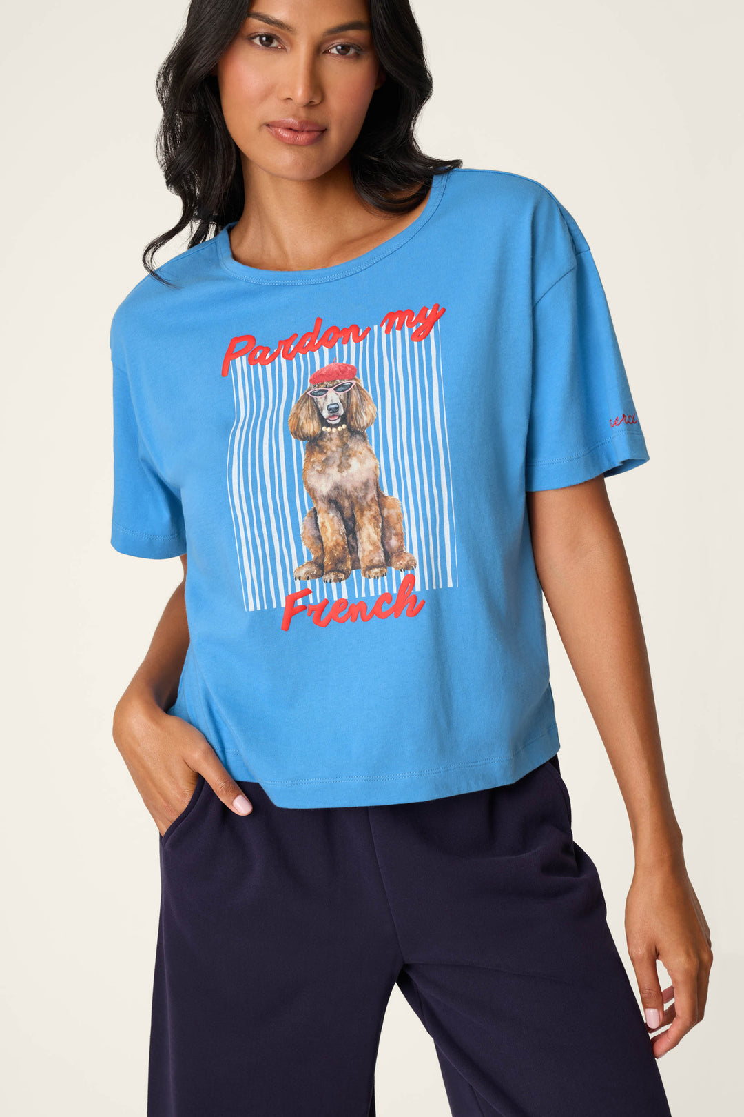 A woman wearing the P.J. Salvage C'est La Vie "Pardon My French" Graphic T-Shirt, featuring an illustration of a dog and red lettering, paired with dark blue pants.