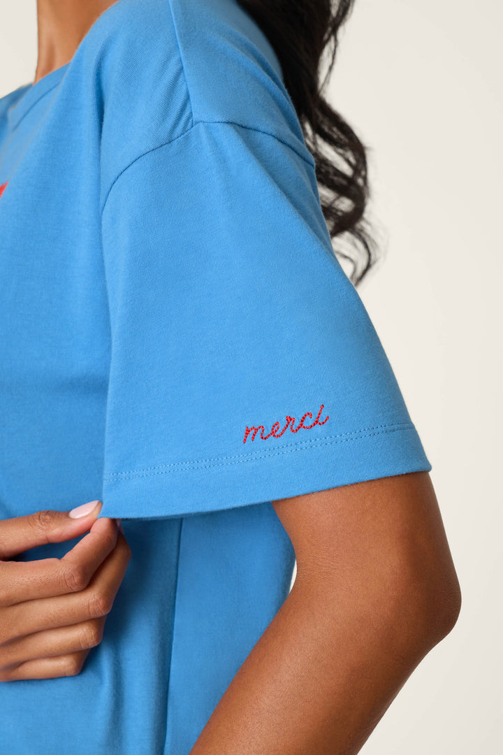 A person wearing the P.J. Salvage C'est La Vie "Pardon My French" Graphic T-Shirt in blue, which features "merci" embroidered in red on the sleeve. The person's hand rests at their side, with their hair visible and a plain, light-colored background behind them.