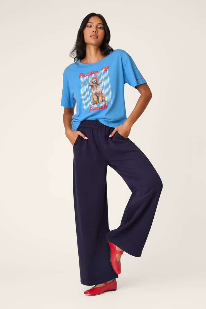 A woman stands against a plain background with one hand in her pocket and one foot slightly raised, wearing the P.J. Salvage C'est La Vie "Pardon My French" Graphic T-Shirt in blue, paired with loose navy blue pants and red flats.