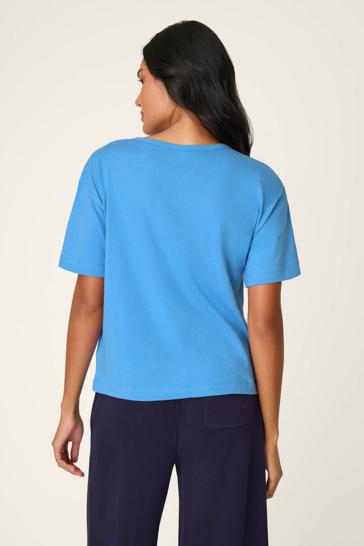 A woman with long dark hair is seen from behind, wearing a P.J. Salvage C'est La Vie "Pardon My French" graphic T-shirt in light blue and dark pants, standing against a plain light background.