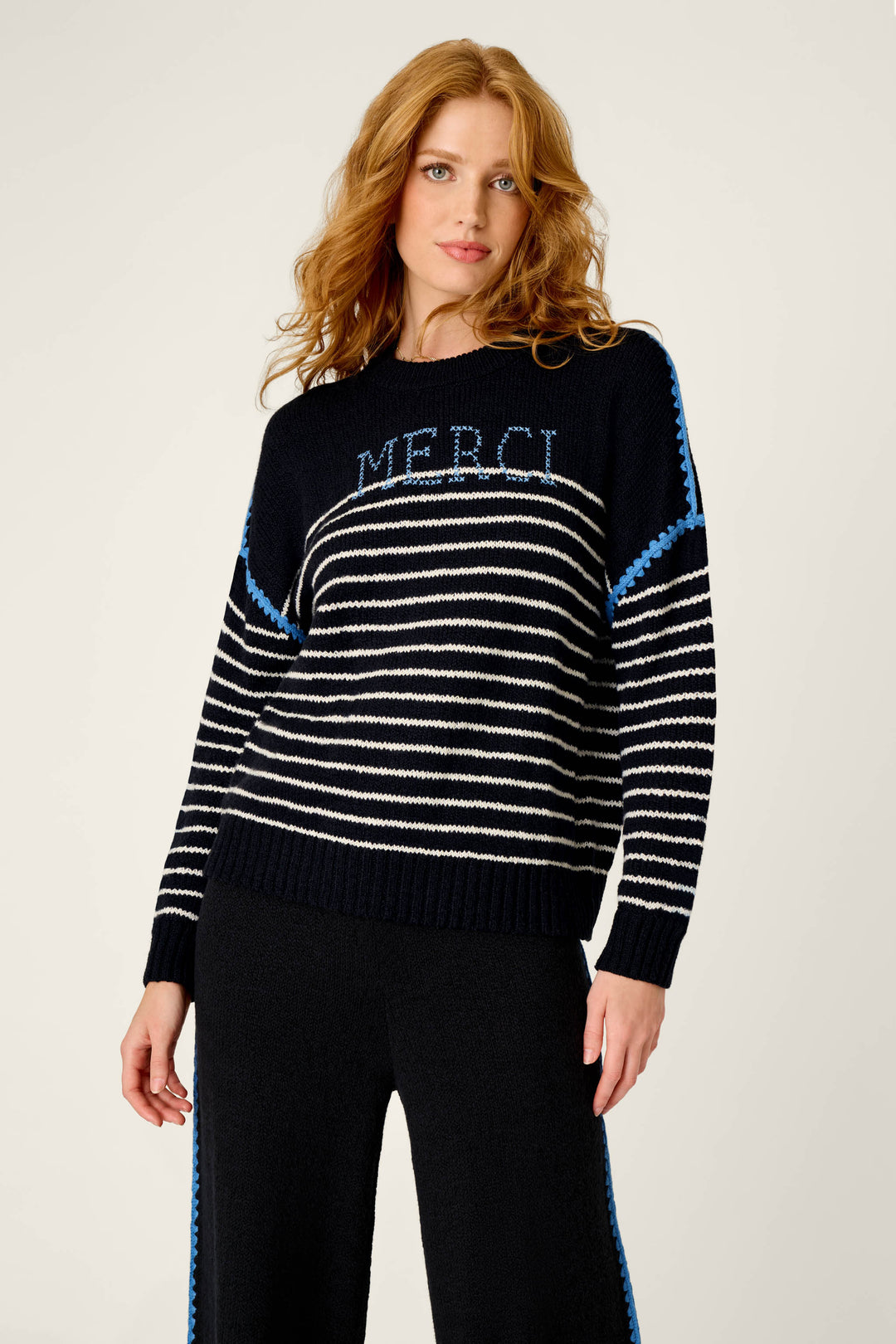 A woman with wavy red hair wears the P.J. Salvage C'est La Vie Merci Sweater—featuring black and white stripes with "MERCI" in blue letters—paired with matching black pants, standing against a plain light background.