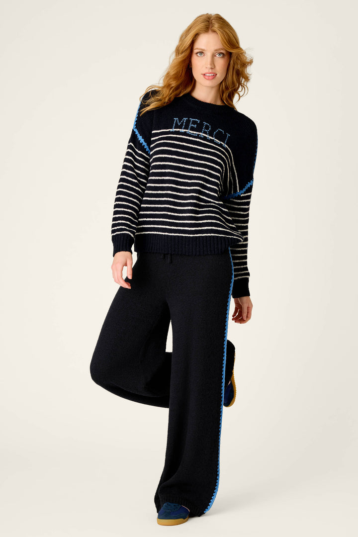 A woman with long, wavy red hair wears the P.J. Salvage C'est La Vie Merci Sweater—a black and white striped top with MERCI text—paired with matching wide-leg pants featuring blue accents, as she stands on one leg against a plain light background.