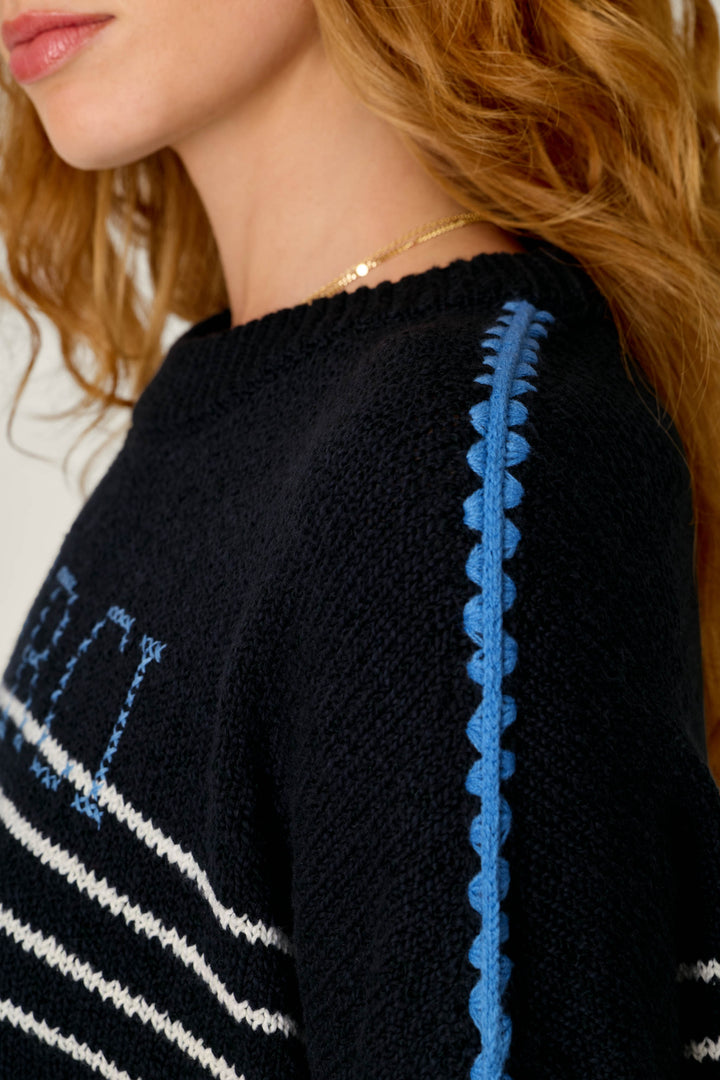 Close-up of a person's side shoulder wearing a black knit sweater with white stripe details and a blue scalloped trim running down the shoulder.