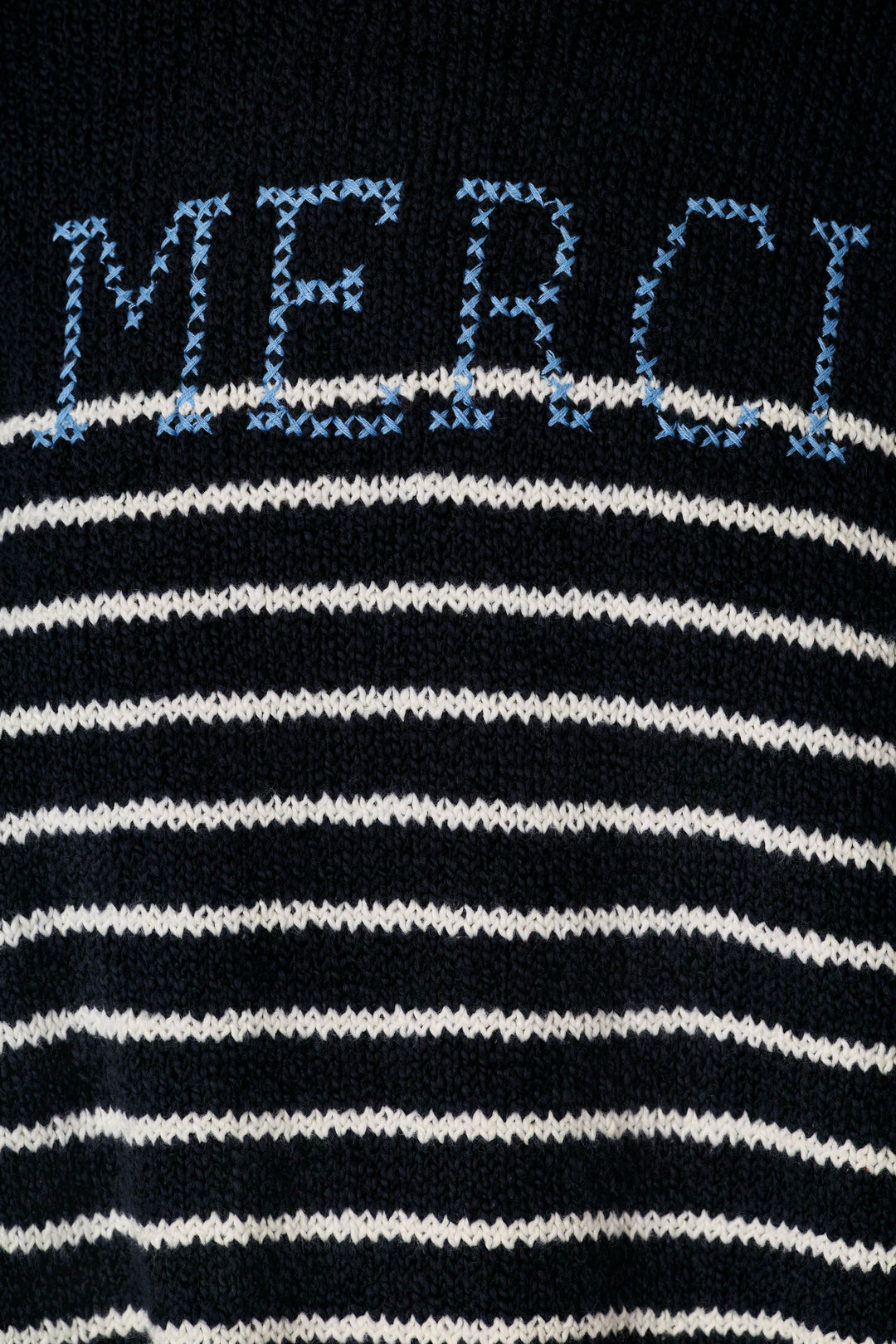 Close-up of a knit sweater featuring horizontal black and white stripes, with the word 'MERCI' knitted across the chest in light blue.