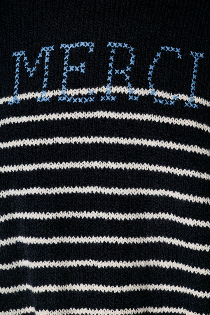 Close-up of a knit sweater featuring horizontal black and white stripes, with the word 'MERCI' knitted across the chest in light blue.