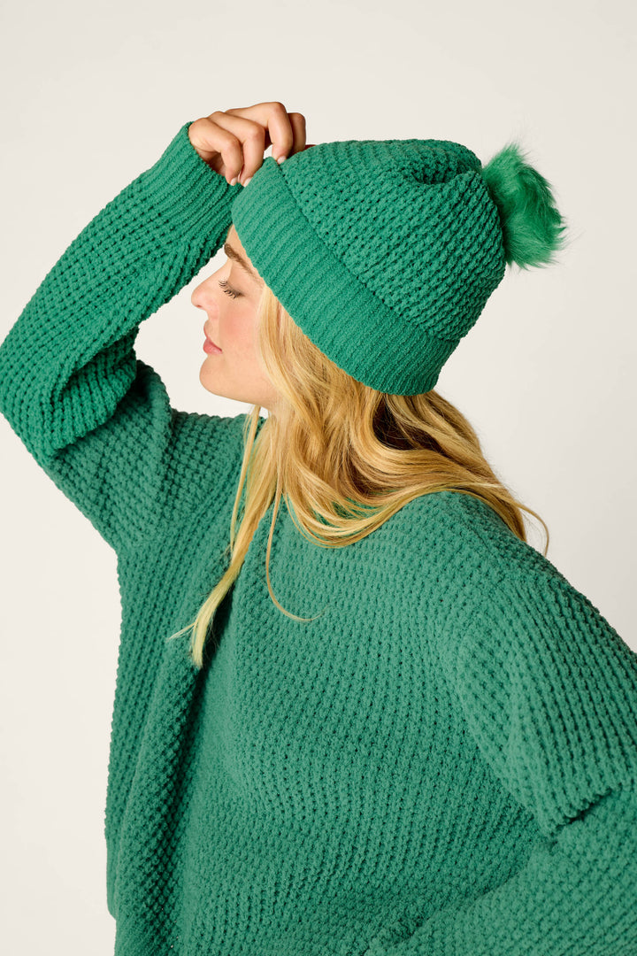 A woman in a textured green sweater tilts her head to the side while lifting the brim of her P.J. Salvage Cozy Chenille Waffle Green Pompom Beanie, set against a plain light background.