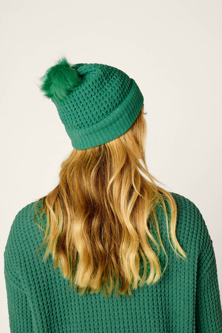 A person with long, wavy blonde hair is seen from behind against a plain background, wearing a P.J. Salvage Cozy Chenille Waffle Green Pompom Beanie paired with a matching green sweater.