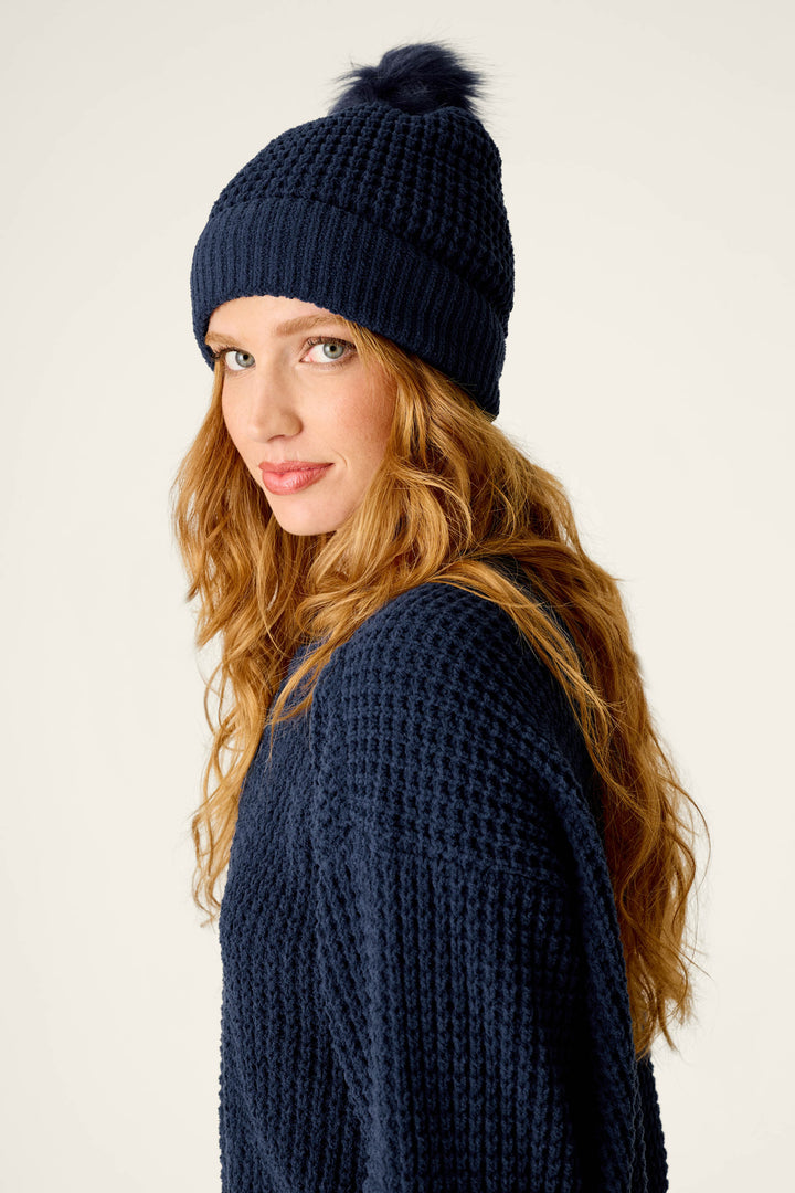 A woman with long red hair wears the P.J. Salvage Cozy Chenille Waffle Deep Navy Pompom Beanie, paired with a navy blue knit sweater, as she looks over her shoulder against a plain light background.