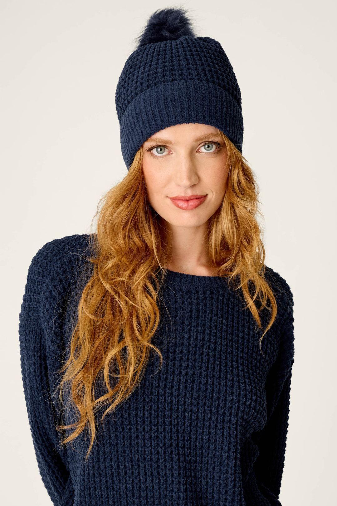 A woman with long wavy red hair is wearing a navy blue knit sweater and the P.J. Salvage Cozy Chenille Waffle Deep Navy Pompom Beanie, looking at the camera against a plain background.