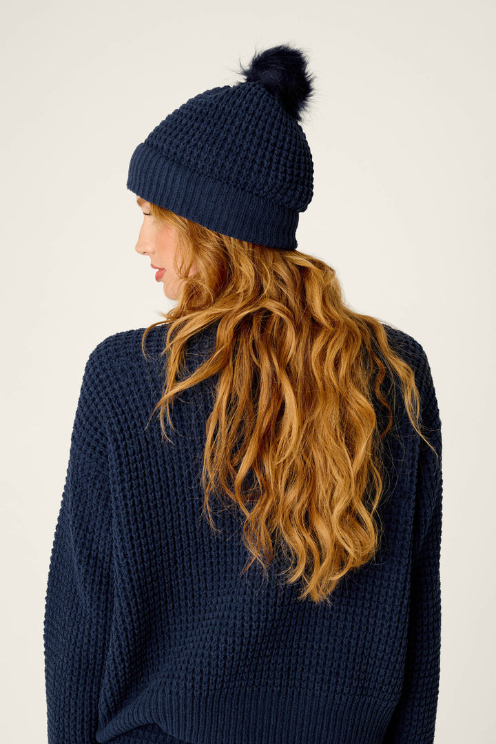 A person with long, wavy auburn hair is seen from behind, wearing a navy blue knit sweater and the P.J. Salvage Cozy Chenille Waffle Deep Navy Pompom Beanie.