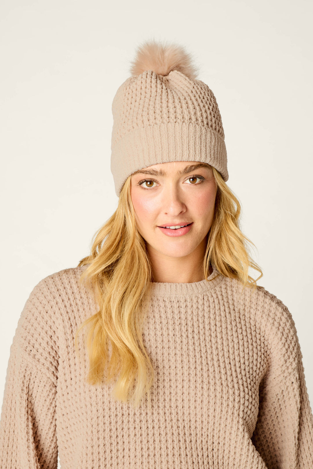 A woman with long blonde hair wears the P.J. Salvage Cozy Chenille Waffle Smoky Beige Pompom Beanie, paired with a beige knit sweater, and smiles softly at the camera against a plain background.