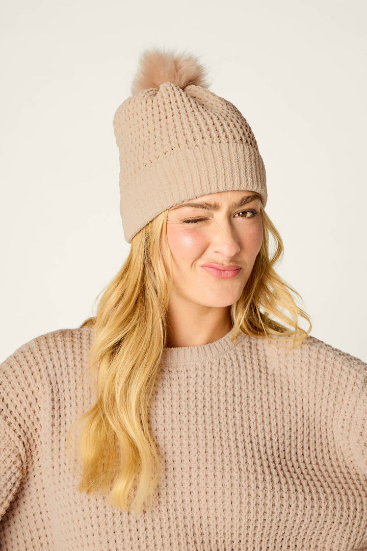 A woman with long blonde hair winks and smiles against a plain background, wearing a beige knit sweater and the P.J. Salvage Cozy Chenille Waffle Smoky Beige Pompom Beanie.