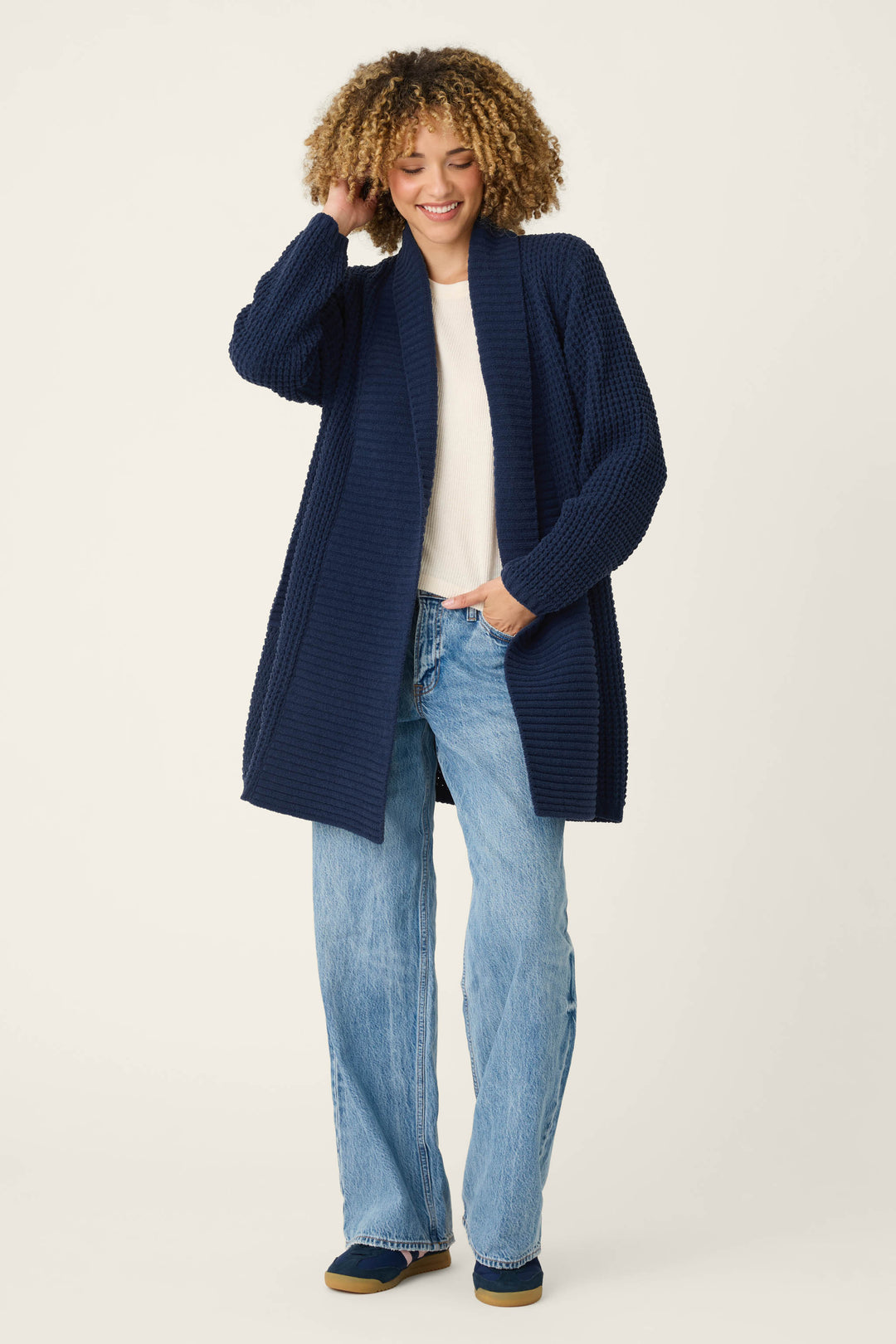 A person with curly hair stands and smiles against a plain background, wearing a P.J. Salvage Cozy Chenille Waffle Deep Navy Cardigan over a white top, paired with light blue wide-leg jeans and navy shoes, with one hand touching their hair.