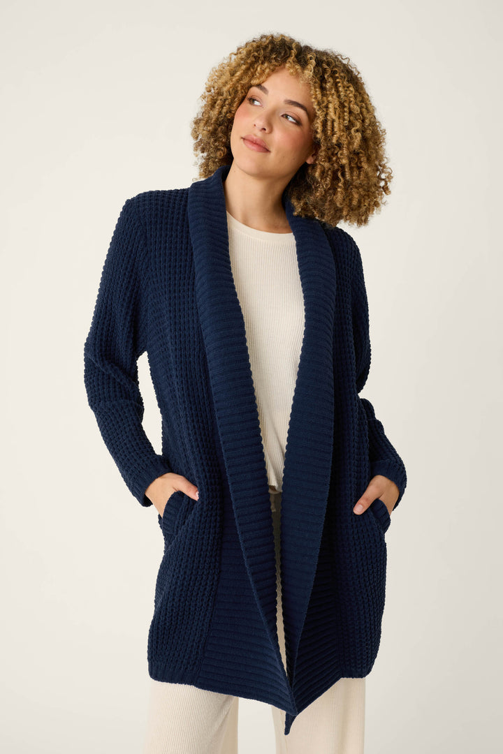 Wearing the P.J. Salvage Cozy Chenille Waffle Deep Navy Cardigan, a person with curly hair stands against a plain background with hands in the cardigan pockets, layering it over a light-colored outfit.