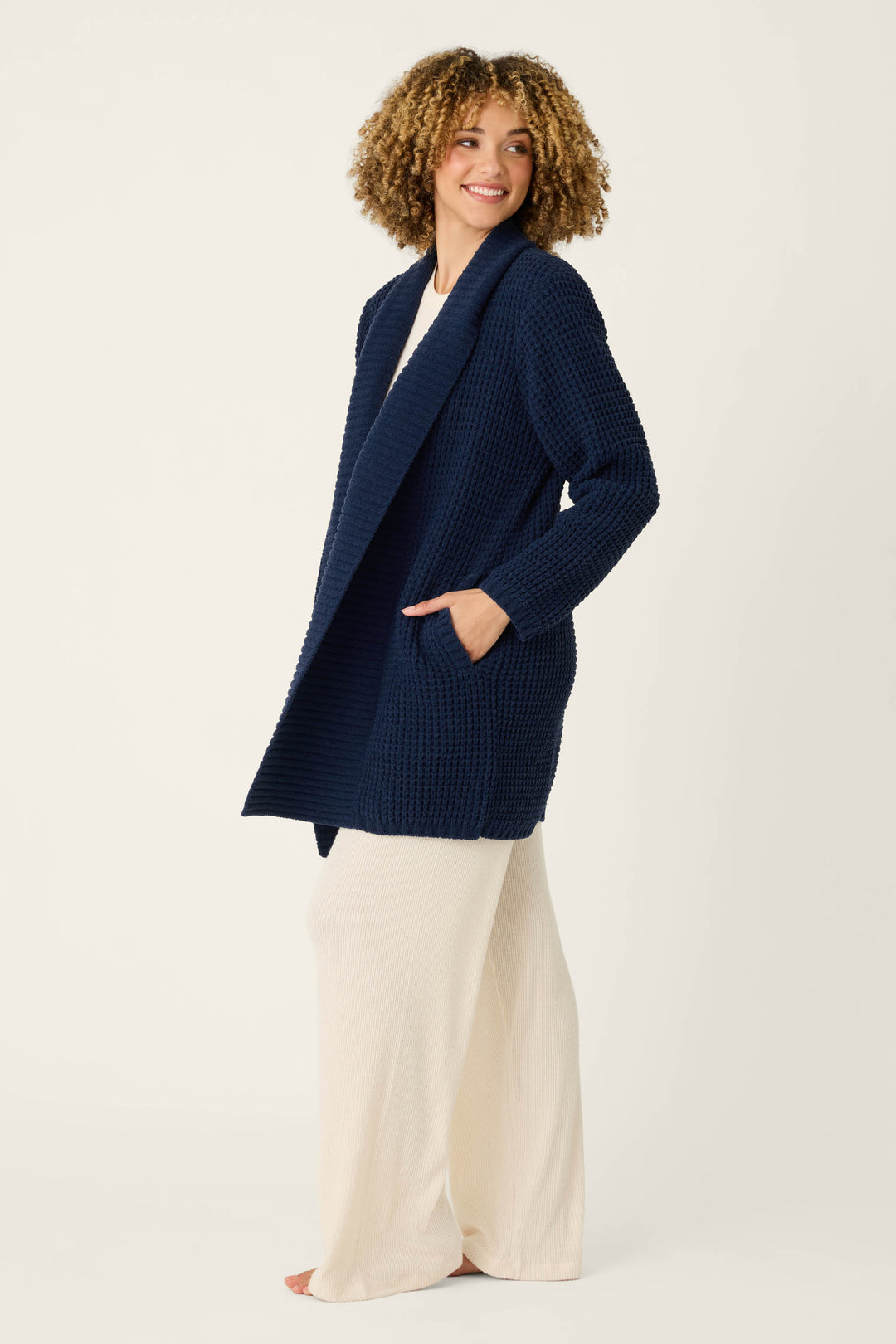 Wearing the P.J. Salvage Cozy Chenille Waffle Deep Navy Cardigan over a long, cream-colored dress, a person with curly hair stands barefoot against a plain light background.