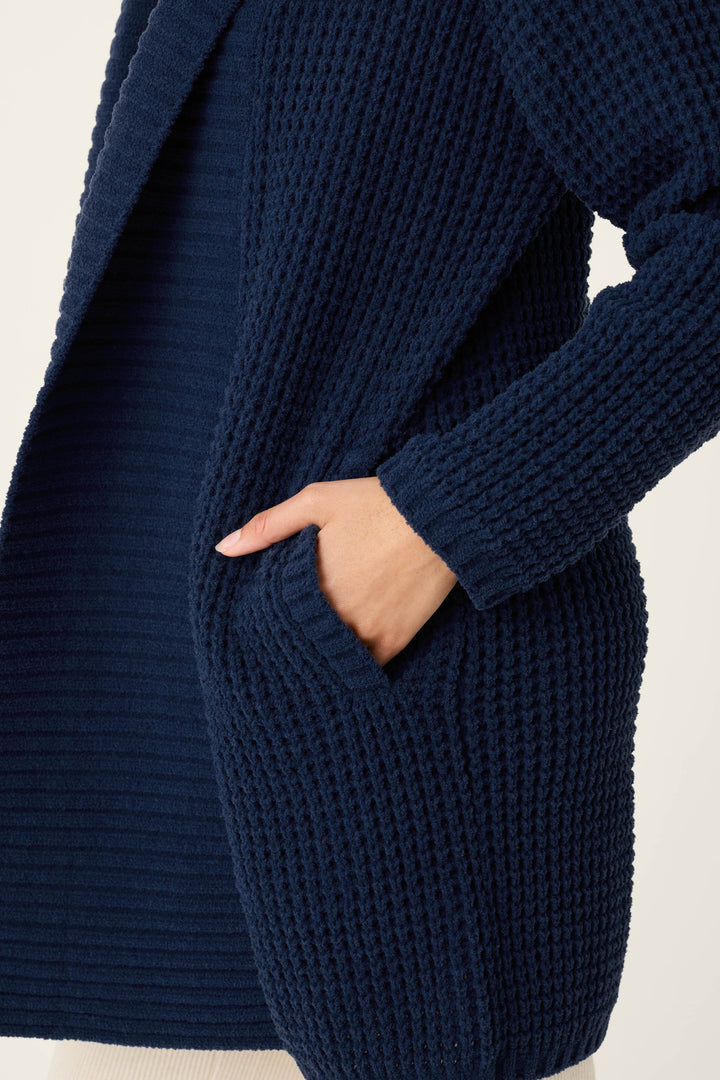 A person is shown from the torso up against a plain light background, wearing the P.J. Salvage Cozy Chenille Waffle Deep Navy Cardigan with their right hand tucked into a pocket.