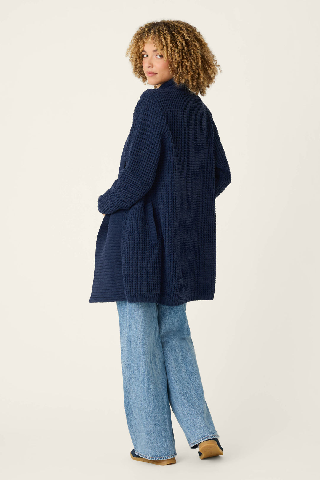 A person with curly hair wears the Cozy Chenille Waffle Deep Navy Cardigan by P.J. Salvage, paired with light blue wide-leg jeans and brown shoes, standing and looking over their shoulder against a plain light background.